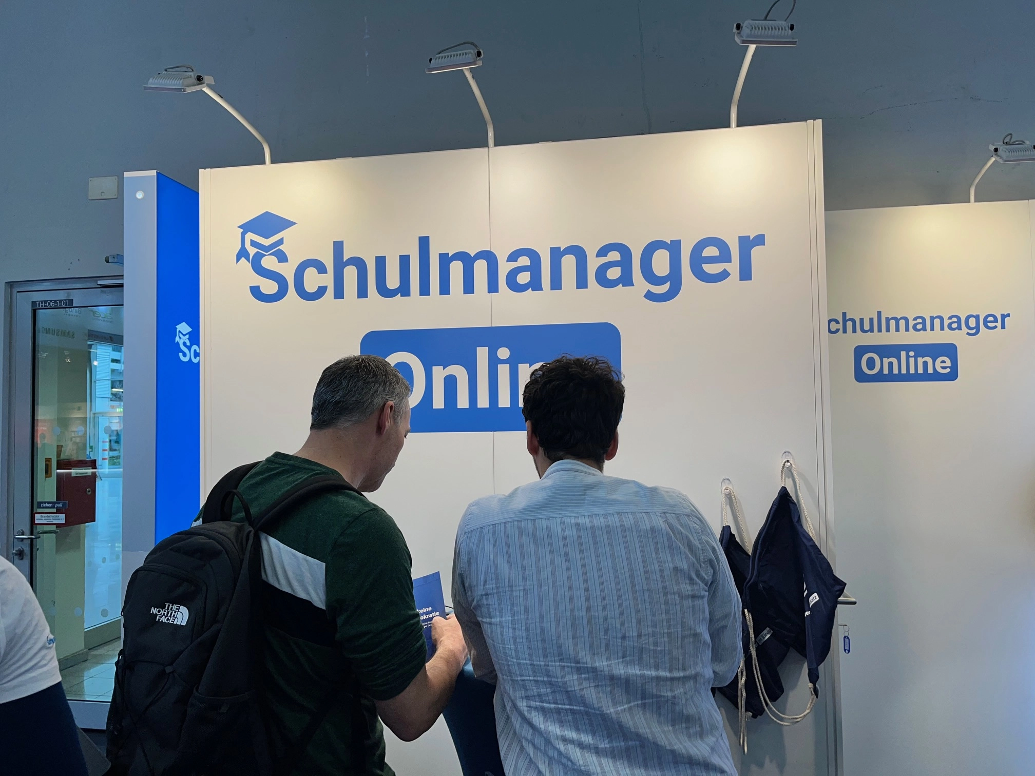 Messestand von Schulmanager Online. Ein großes blaues Schulmanager-Online Logo ist im Hintergrund zu sehen. Davor stehen ein Mitarbeiter von Schulmanager Online, der im Gespräch mit einem Interessenten.